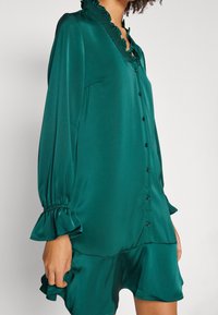 Blusa in satin turchese con collo a volant, maniche lunghe a sbuffo e chiusura a bottoni. Presenta un orlo a strati e una consistenza liscia.