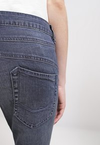 Persona vistiendo jeans azul oscuro con el bolsillo trasero visible, de pie contra un fondo claro y liso, con el brazo relajado a su lado.