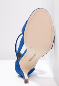 Scarpa con tacco alto in suede blu, con cinturino alla caviglia, suola beige, taglia 39, nome del marchio "Tamaris" visibile sulla suola, inclinata su sfondo bianco.