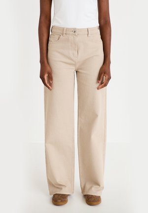 Persoon draagt beige wijde broekspijpen, bruine schoenen en een witte ingestoken top, staand tegen een effen lichte achtergrond.