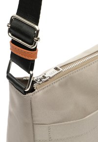 Beige Stofftasche mit strukturierter Oberfläche, schwarzem verstellbarem Tragegurt mit silbernen Beschlägen und einem Reißverschluss mit metallischen Details.