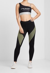 Leggings de sport noirs et verts olive avec une coupe ajustée, associés à un crop top noir et gris. Présente un logo bien visible sur le haut.