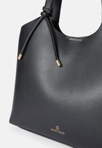 Μαύρη δερμάτινη τσάντα tote με λεία υφή, καμπυλωτό σχήμα και κόμπο στη λαβή. Διαθέτει χρυσές λεπτομέρειες και ορατό λογότυπο μάρκας.