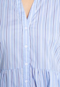 Lichtblauwe blouse met verticale witte en marineblauwe strepen, voorzien van een V-hals, knoopsluiting en een peplumzoom. Zacht katoenen stof.