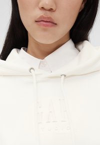 Hvit hettegenser med et strukturert logo foran, med snørehette og en krage fra en underliggende hvit skjorte.