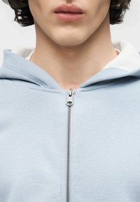 Νεαρό άτομο που φοράει ανοιχτό μπλε φούτερ με φερμουάρ κλειστό μέχρι πάνω, εμφανίζοντας τον λαιμό και το κάτω μέρος του προσώπου.