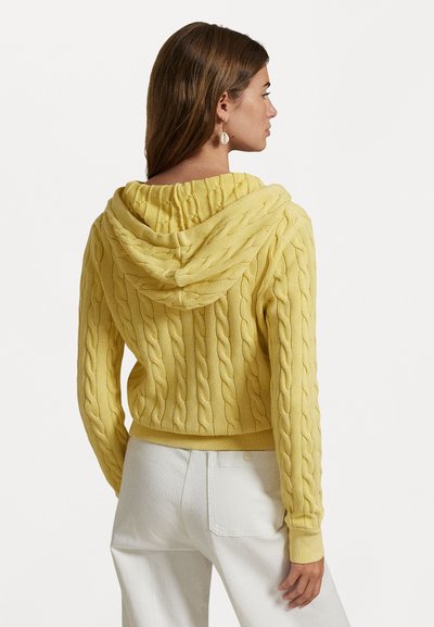 Gele kabelgebreide hoodie met een ontspannen pasvorm, voorzien van een opvallende kap en geribde boorden, gecombineerd met witte high-waisted broeken.