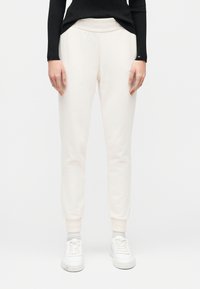 Γυναίκα που φοράει μπεζ παντελόνια jogger, λευκά αθλητικά παπούτσια και μαύρο μακρυμάνικο τοπ, στέκεται μπροστά σε απλό λευκό φόντο.