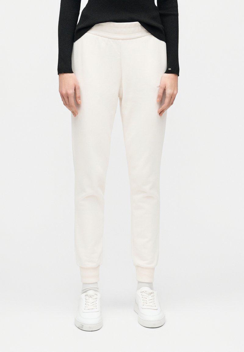 Γυναίκα που φοράει μπεζ παντελόνια jogger, λευκά αθλητικά παπούτσια και μαύρο μακρυμάνικο τοπ, στέκεται μπροστά σε απλό λευκό φόντο.