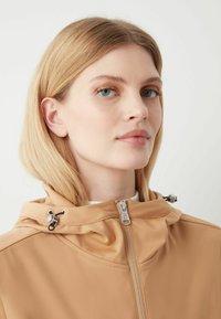 Beige Jacke mit hohem Kragen, Reißverschluss und verstellbaren Kordeldetails in der Nähe der Kapuze. Glatter Stoff mit matter Oberfläche.