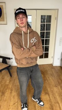 Noor mees, kes kannab pruuni kapuutsiga pusa, musta nokamütsi, millel on kiri "METAL GEAR", halli teksapükse ja must-valgeid tossusid, seisab siseruumis, käed rüpes.