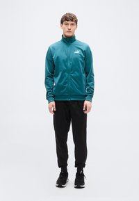 Τζελ εξωτερικό αθλητικό μπουφάν με φερμουάρ και ψηλό γιακά, συνδυασμένο με μαύρο παντελόνι jogging. Το μοντέλο στέκεται μπροστά σε απλό λευκό φόντο.