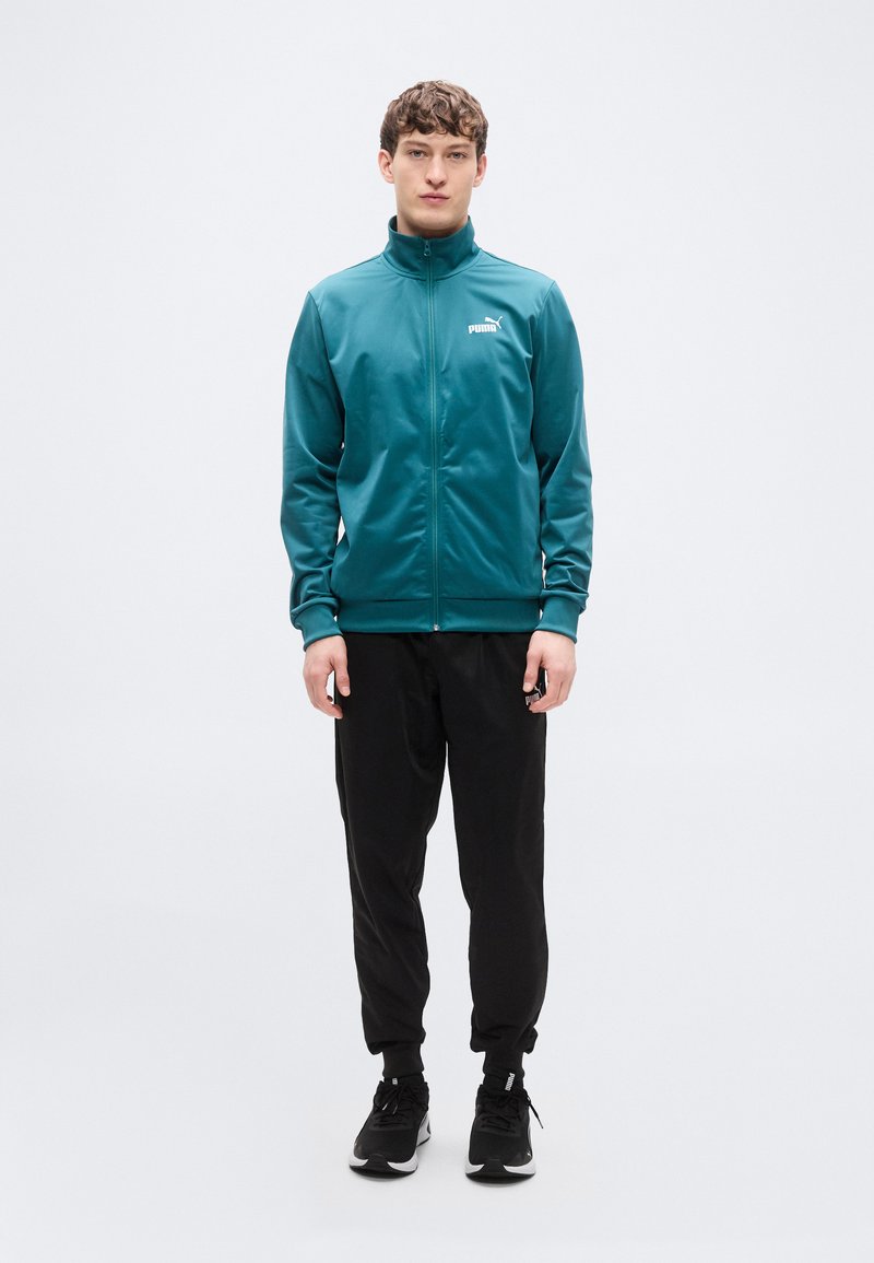 Τζελ εξωτερικό αθλητικό μπουφάν με φερμουάρ και ψηλό γιακά, συνδυασμένο με μαύρο παντελόνι jogging. Το μοντέλο στέκεται μπροστά σε απλό λευκό φόντο.