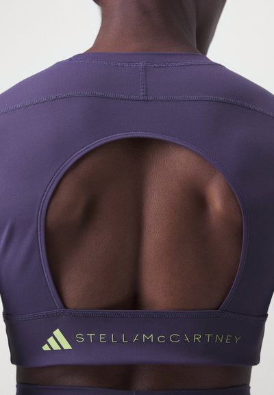 Fialový športový crop top s prestrihom na chrbte, so bezšvovou textúrou a zeleným logom "STELLA McCARTNEY" na dolnej strane.