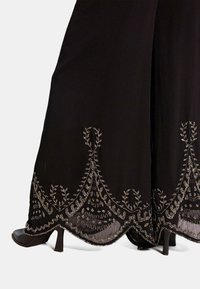Pantaloni neri a gamba larga con un intricato ricamo argentato all'orlo, dotati di una texture fluida e una leggera lucentezza. Scarpe nere con tacco visibili.