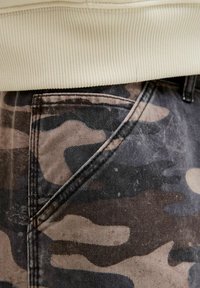 Jean camouflage avec des motifs noirs, marron et beiges présentant une surface texturée. La taille est en matériau côtelé contrastant.