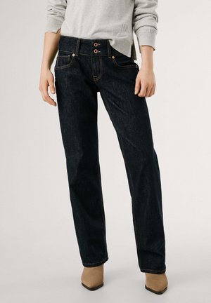 Donkerblauwe denim jeans met twee knoopsluitingen, vijf zakken en contrasterende gouden stiksels, gecombineerd met beige haklaarzen.