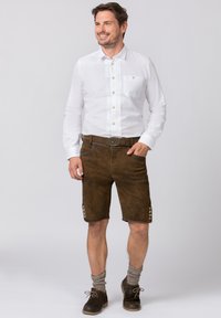 Witte overhemd met plooien, gecombineerd met bruine leren shorts met decoratieve knopen. Bruine sokken en schoenen maken de outfit compleet.