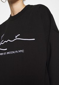 Acercamiento a una sudadera negra con un logo en cursiva blanca y el texto "1989 EST. BROOKLYN, NYC", llevada por una persona con aros en las orejas.