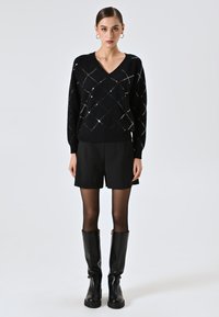 Maglione nero con scollo a V, decorato con motivo a rete di diamanti in paillettes argentee. Abbinato a pantaloncini neri e stivali neri sopra il ginocchio.