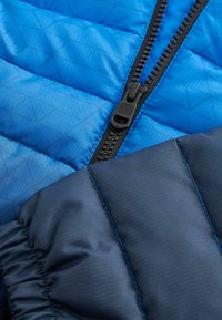 Tissu de veste rembourrée bleue avec des manches contrastantes navy, featuring une fermeture éclair noire et une texture géométrique. Design lisse et isolé.