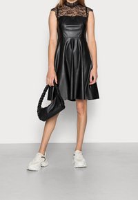 Schwarzes Kunstlederkleid mit einer Spitzen-Bodice, A-Linien-Rock, hohem Kragen, kombiniert mit einer geflochtenen schwarzen Tasche und klobigen weißen Sneakers.