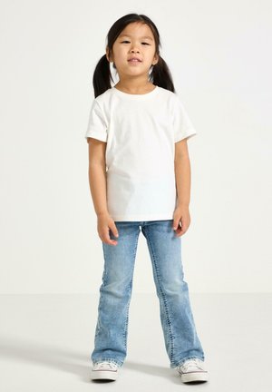 Biała bawełniana koszulka z krótkim rękawem, zestawiona z jasnoniebieskimi rozkloszowanymi dżinsami. Sneakersy z białą cholewką z płótna i gumowymi podeszwami.