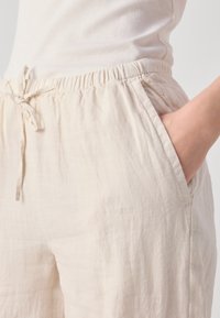 Personne portant un pantalon en lin beige avec une taille élastique et un cordon de serrage, main placée dans la poche latérale, associé à un haut blanc côtelé.