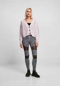 Lys lilla strikket cardigan med store knapper, sammen med svarte og hvite mønstrede leggings med mesh-utskjæringer. Svarte joggesko på.