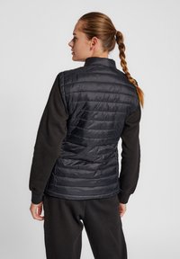 Svart pufferväst med kviltat mönster, hög krage och ärmlös design, tillsammans med en mörk långärmad skjorta. Slät textur.