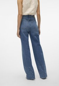 Jeans blu a vita alta e gamba larga con una texture di denim liscia. Presentano un orlo frangiato ornato da dettagli in perle bianche.