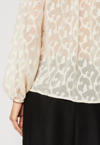 Blouse en crème légère et transparente avec un motif texturé blanc ; manches bouffantes avec des poignets élastiques ; l'ourlet est au-dessus d'une jupe noire.