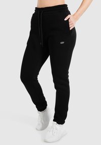 Schwarze Jogginghosen aus weichem Stoff, mit einem Kordelzugbund, seitlichen Taschen und einem kleinen Logo. Kombiniert mit weißen Sportschuhen.