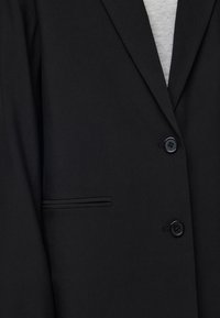 Gros plan sur une veste noire avec deux boutons noirs, un revers étroit, une poche passepoilée et une chemise gris clair visible en dessous.