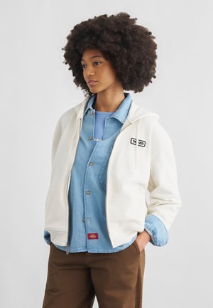 Γυναίκα με άφρο χτένισμα που φοράει λευκή φούτερ με φερμουάρ πάνω από μπλε τζιν πουκάμισο και καφέ παντελόνι, στέκεται με τα χέρια στις τσέπες.