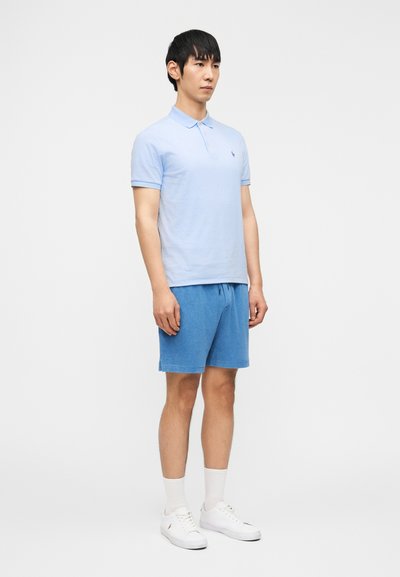 Νέος άνδρας στέκεται φορώντας ανοιχτό μπλε πόλο πουκάμισο, μπλε σορτς με κορδόνι, λευκές κάλτσες και λευκά αθλητικά παπούτσια μπροστά σε απλό φόντο.