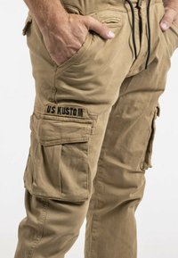 Pantalons cargo beiges avec une taille à cordon, de multiples poches et un logo "US KUSTOM" visible sur le côté. Matière douce et légère.