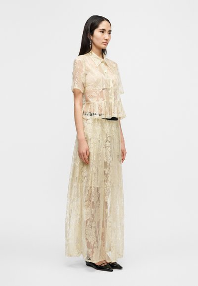 Τοπ από δαντέλα και μακριά φούστα σε απαλό μπεζ, με floral σχέδια, κοντά μανίκια, σχεδιασμό πεπλούμ και μαύρη μέση. Μαύρα μυτερά παπούτσια.