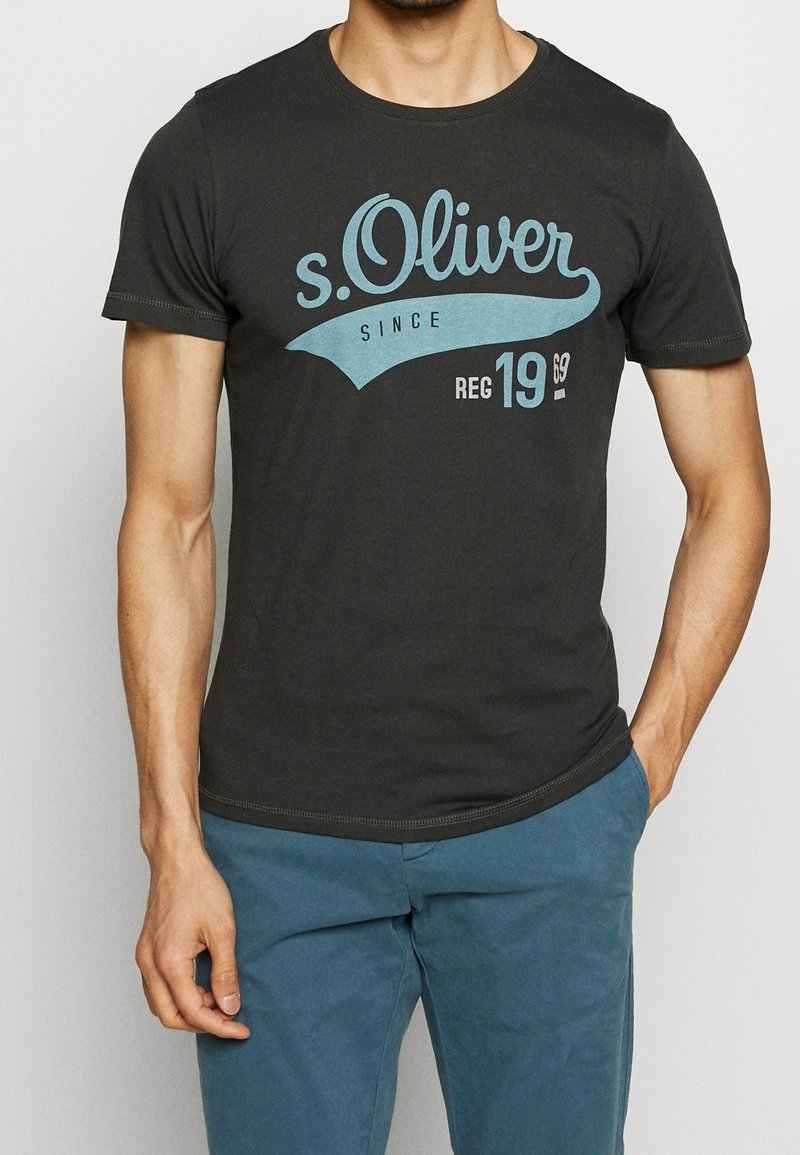 Homme portant un t-shirt noir à manches courtes avec le texte bleu "s.Oliver Since Reg 1969" et un pantalon bleu, debout une main dans la poche.