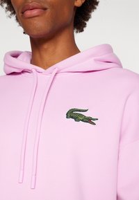 Felpa rosa realizzata in un materiale morbido, con un logo di coccodrillo verde ricamato sul lato sinistro del petto e coulisse regolabili.