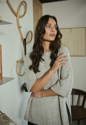 Light gray textured sweater with short sleeves and side slits, worn by a person with long, wavy brown hair in a minimalist setting.