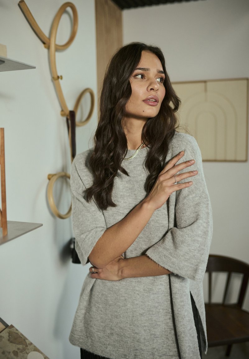 Pull en maille texturée gris clair avec des manches courtes et des fentes sur les côtés, porté par une personne aux longs cheveux bruns ondulés dans un cadre minimaliste.
