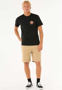 Camiseta negra de manga corta con un colorido logo redondo en el pecho, combinada con pantalones cortos caqui, calcetines negros y zapatillas con cordones blancos.