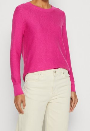 Personne portant un pull texturé à manches longues rose vif et un pantalon taille haute écru sur un fond uni.