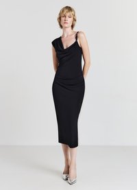 Schwarzes, figurbetontes Kleid mit drapiertem Ausschnitt und einem Schulterdesign. Glatte Stoffqualität, knielang, akzentuiert durch einen silbernen Riemen an der Schulter.