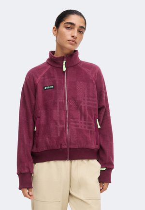 Persona que lleva una chaqueta de forro polar granate con tiradores verdes en la cremallera y pantalones beige, de pie frente a un fondo gris claro liso.