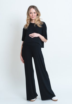 Schwarzes, kurzes Oberteil mit weiten Ärmeln, kombiniert mit lockeren schwarzen Hosen. Das Model trägt hellfarbige Schuhe. Glattes Material und lässige Passform.