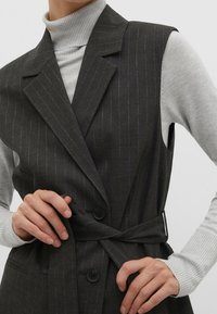 Gilet a righe grigio scuro con design avvolgente, dotato di un fiocco in vita e bottoni neri. Indossato sopra un dolcevita a coste grigio chiaro.