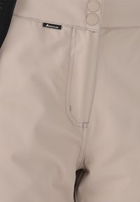 Beige waterdichte broek met een textuurafwerking, voorzien van twee knoopsluitingen en een klein logo-label. De stof lijkt duurzaam en glad.