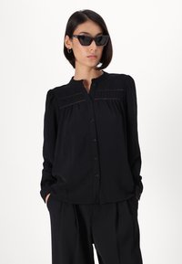 Black button-up shirt with a round collar, pleated detailing on the front, and long sleeves. Smooth texture and relaxed fit.